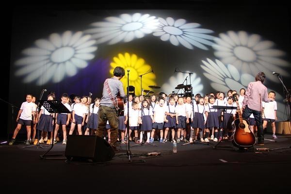舞台の上で「ちめいど」と子供達が歌い、その後ろでお花の映像が映し出されていて黄色の花だけ色がついている写真